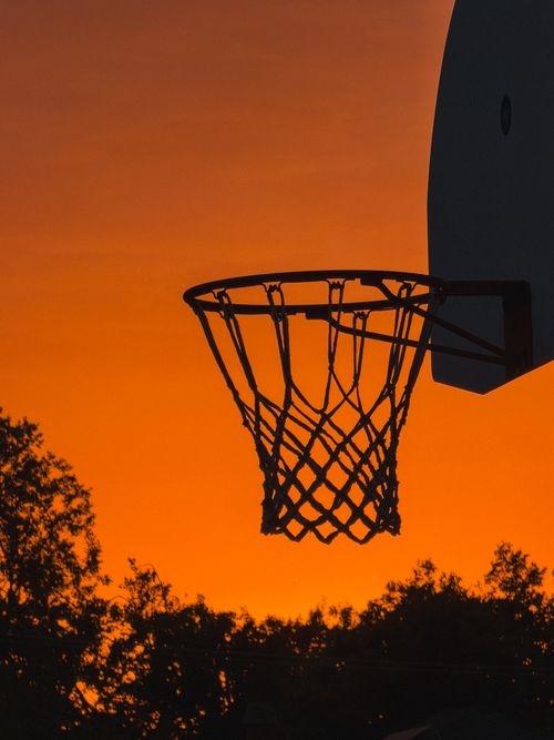 Free A basketball hoop silhouetted against a vivid sunset with trees below, capturing an outdoor sports theme. Stock Photo
