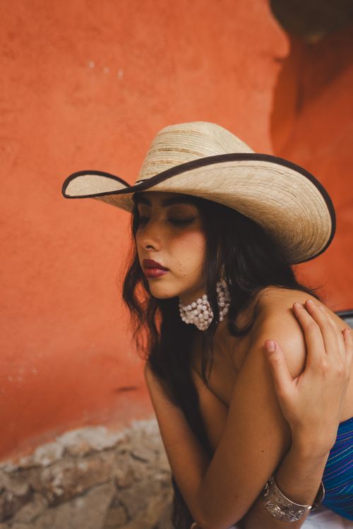 Free Portrait of a woman in a straw hat and earrings, set against a vibrant terracotta wall. Stock Photo