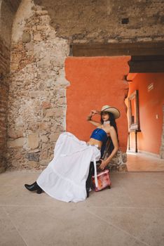Fashionable woman poses in a rustic interior with stone walls, wearing a hat and flowing dress.
