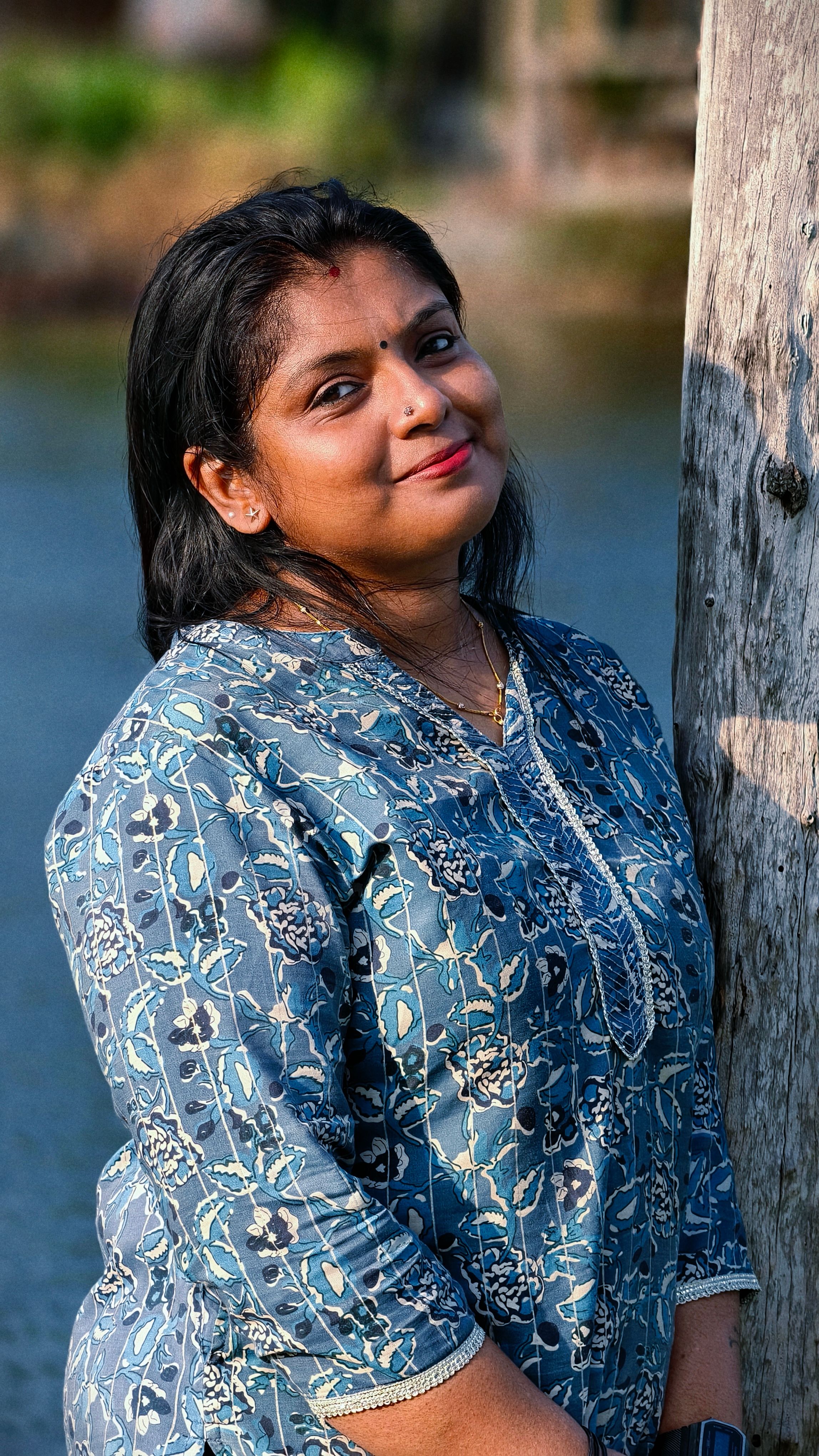 Elegant Indian woman in traditional attire, leaning on a tree by a lake in a serene setting.