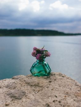 Purple wildflowers in a green glass bottle on a rock by a serene lake under cloudy skies.