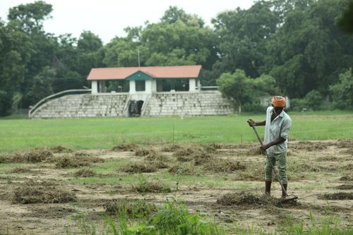 Free A farmer in Raipur, India, working with a tool in a field near a rural building, surrounded by lush greenery. Stock Photo