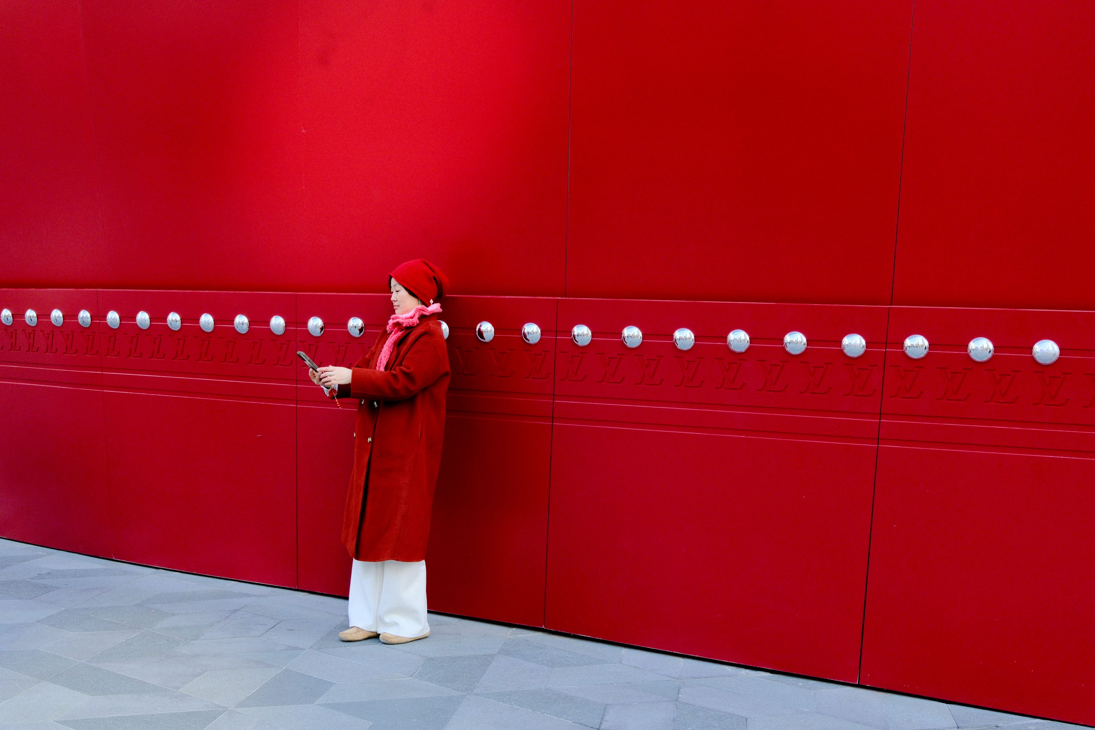 Stylish woman in red coat texting on smartphone against a modern geometric red wall.