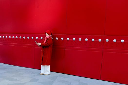 Stylish woman in red coat texting on smartphone against a modern geometric red wall.