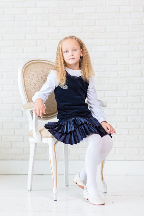 Girl Wearing Blue Dress With White Long Socks Sitting on White Wooden Armchair Beside Wall