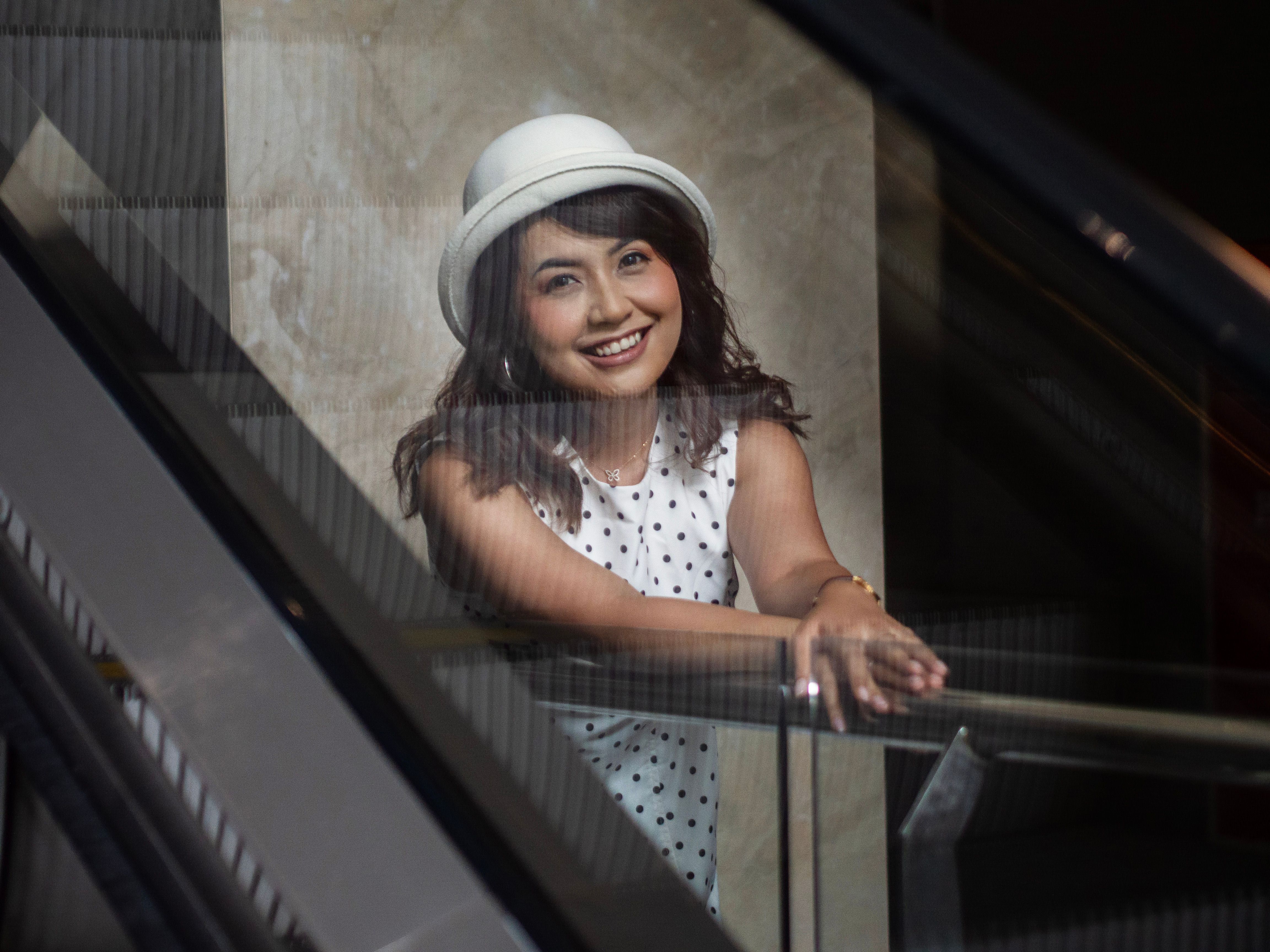 A cheerful woman in a white hat smiles while leaning on an escalator railing.