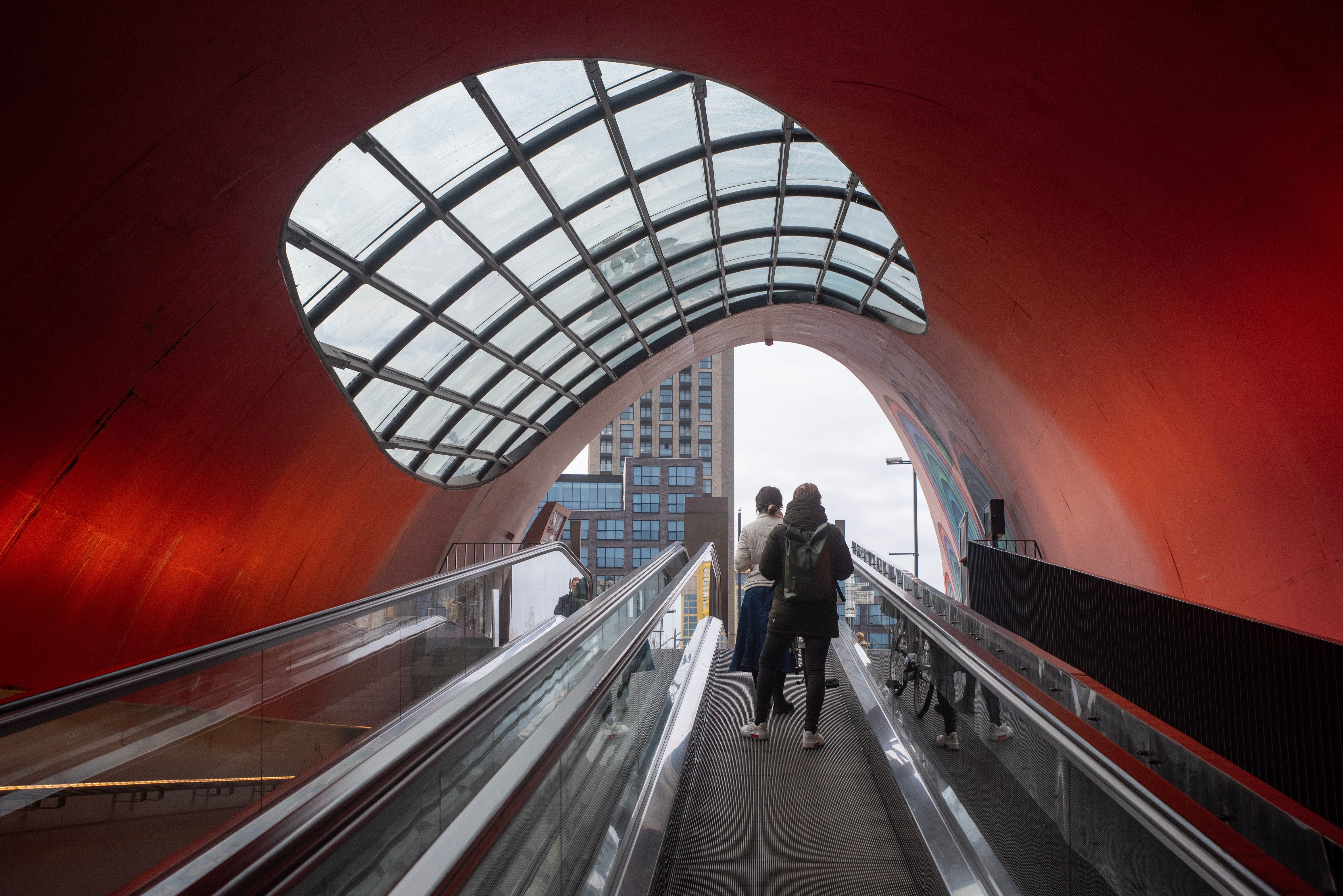 Two people on an escalator in a modern tunnel with red walls and glass roof in Eindhoven.