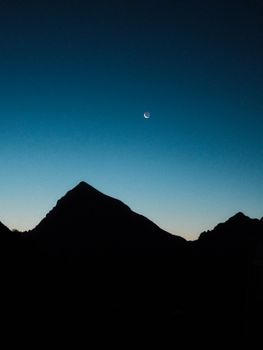 A serene silhouette of a mountain against a clear twilight sky with a crescent moon.