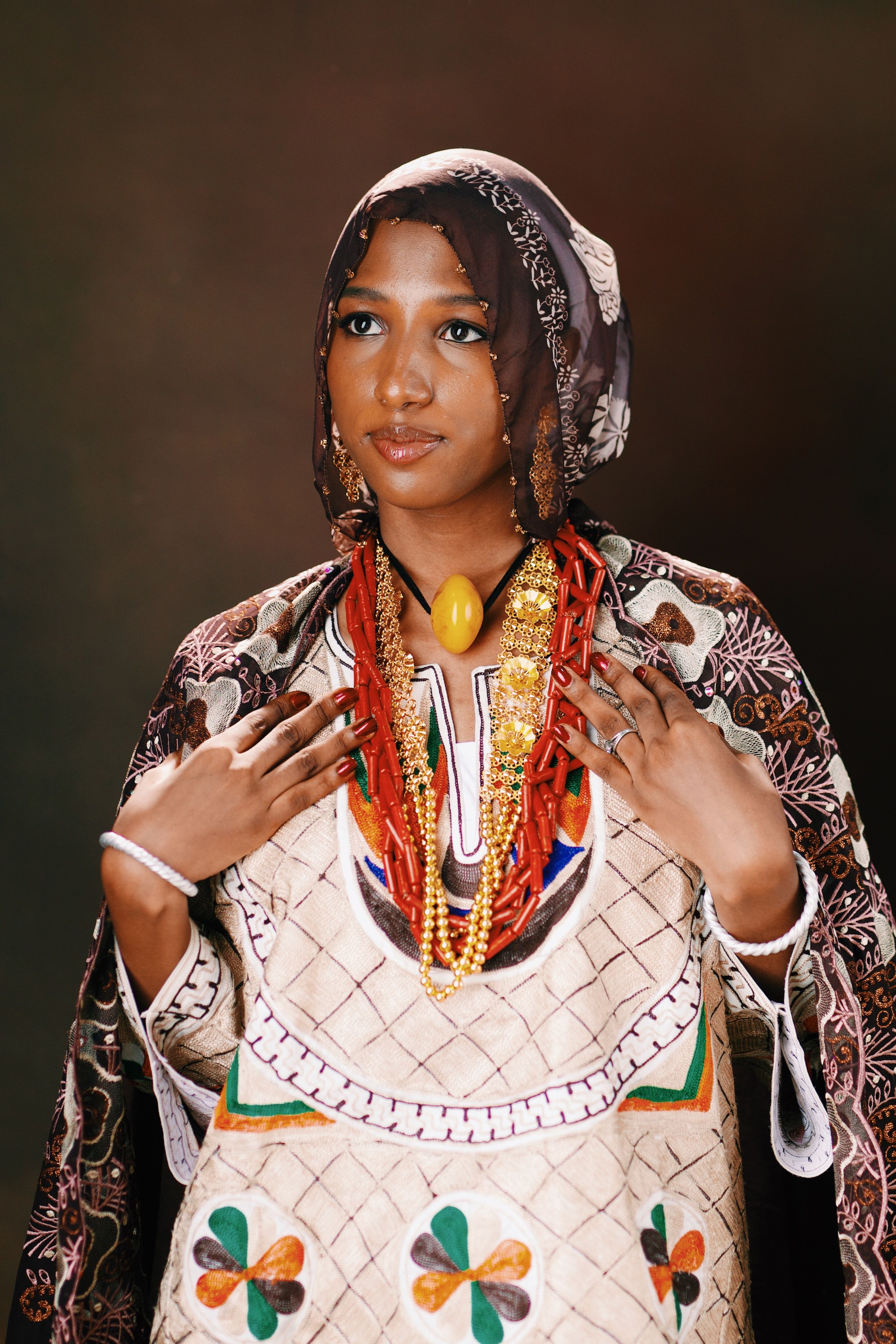 Woman in colorful traditional African dress with intricate jewelry on a neutral background.