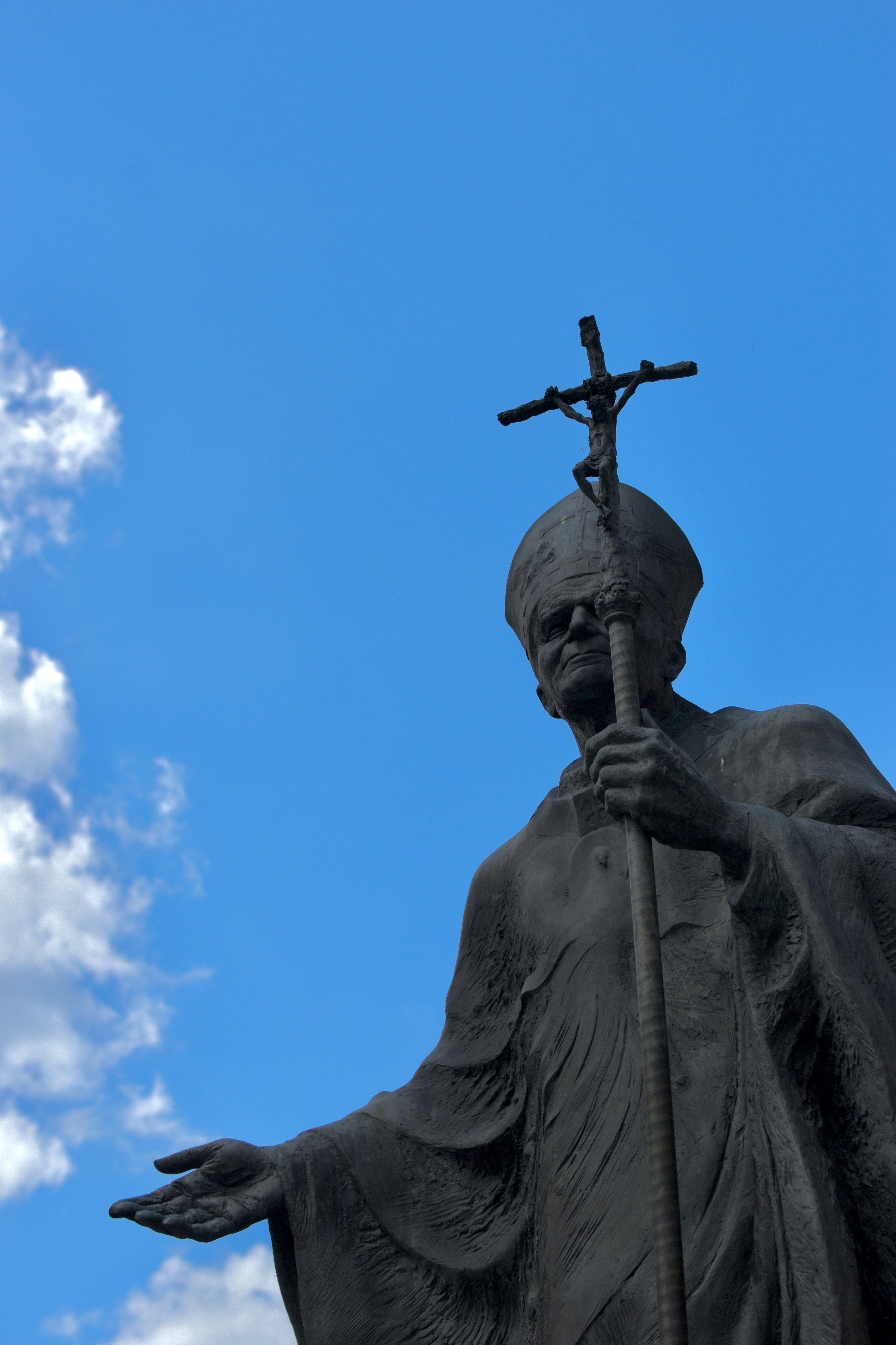 Dramatic statue of a pope holding a cross with a bright blue sky background.