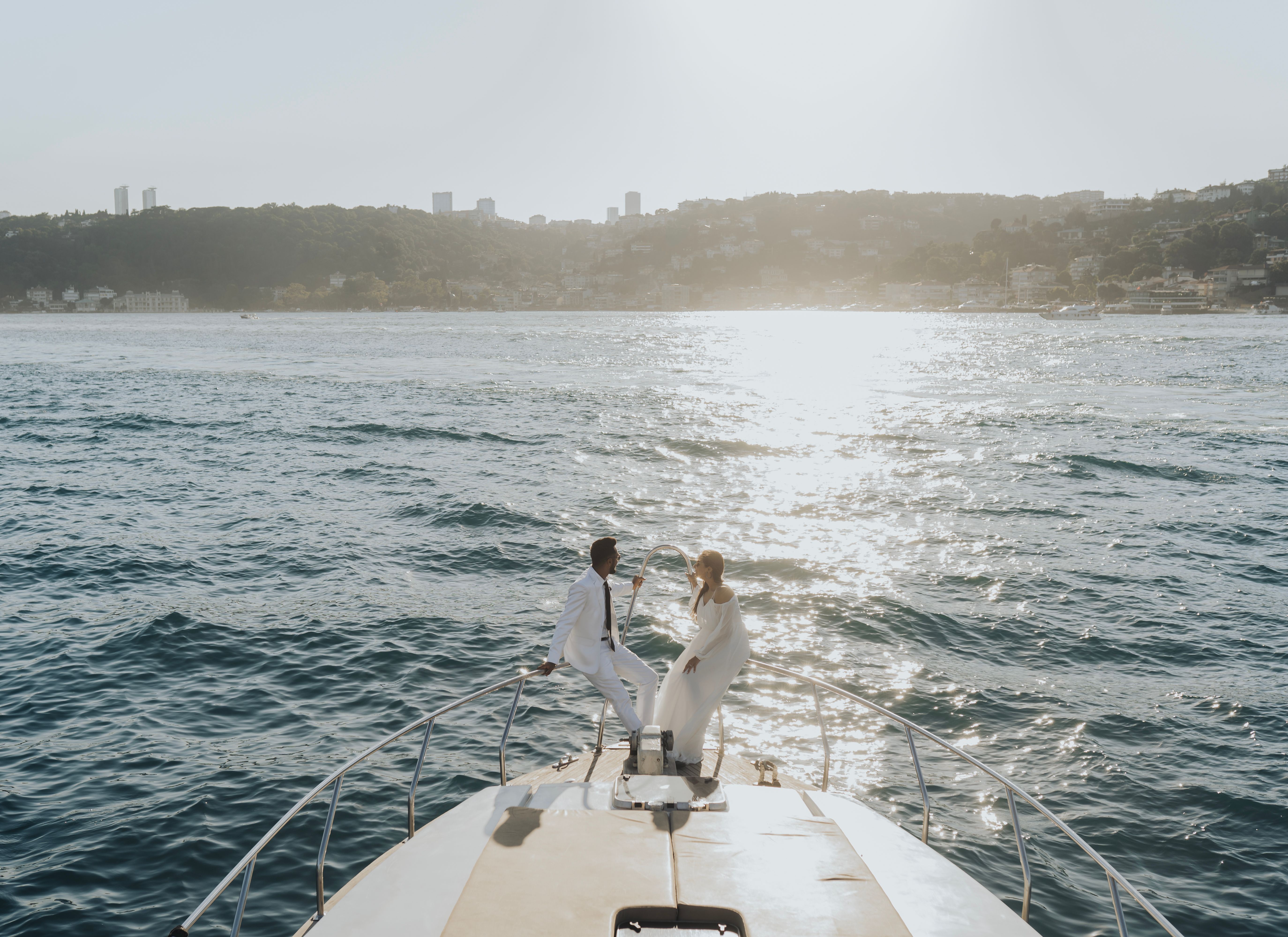 A bride and groom share a romantic moment on a yacht against a serene waterfront backdrop during sunset.