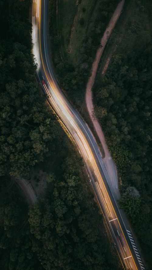 Free Captivating aerial view of a winding road with car light trails through a dense forest at night. Stock Photo
