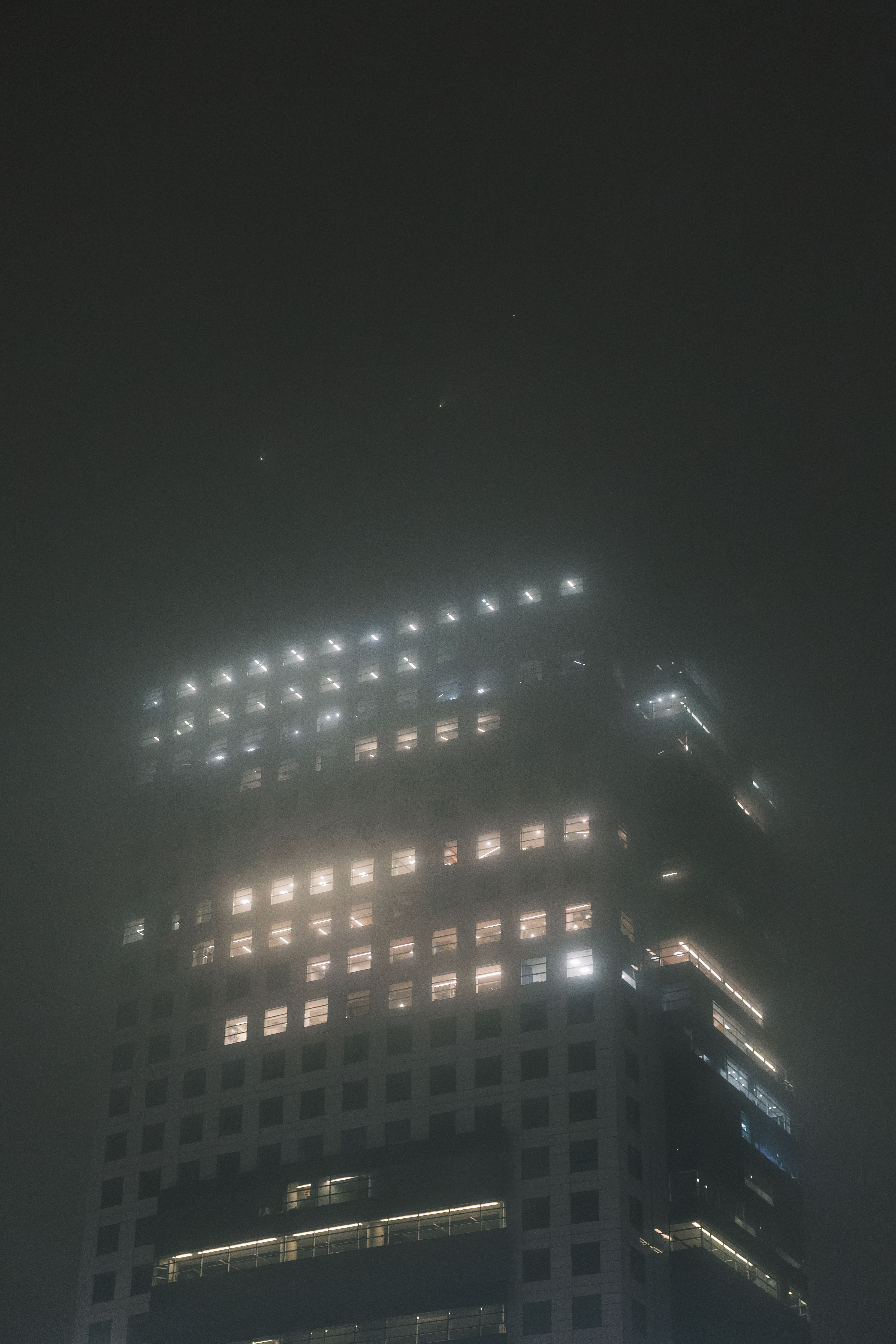 A skyscraper emerges from the fog with glowing windows during a misty night.