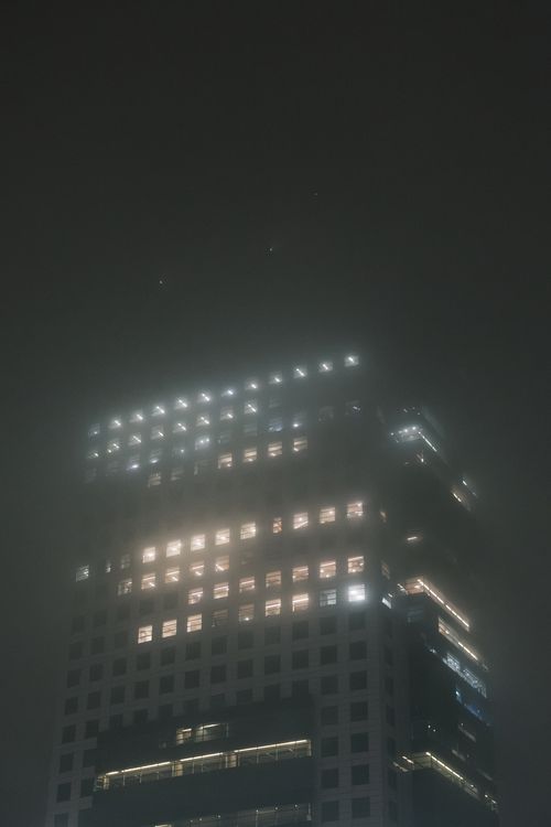 Free A skyscraper emerges from the fog with glowing windows during a misty night. Stock Photo