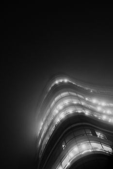 A sleek, modern building with bright lights pierces the dense nighttime fog.