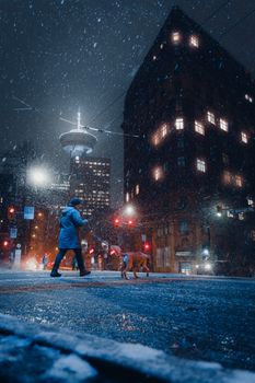 A person walks their dog through a snowy Vancouver street at night, with city lights illuminating the scene.