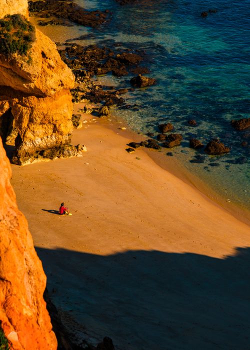 Free A lone person sits on a sandy beach at sunset beside rugged cliffs and blue ocean waters. Stock Photo