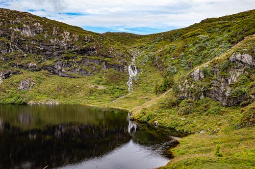 Free Lush green Norwegian mountain landscape with a waterfall cascading into a serene lake under a cloudy sky. Stock Photo