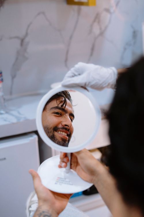 Free A man smiles while viewing himself in a handheld mirror in a salon setting, showcasing satisfaction. Stock Photo