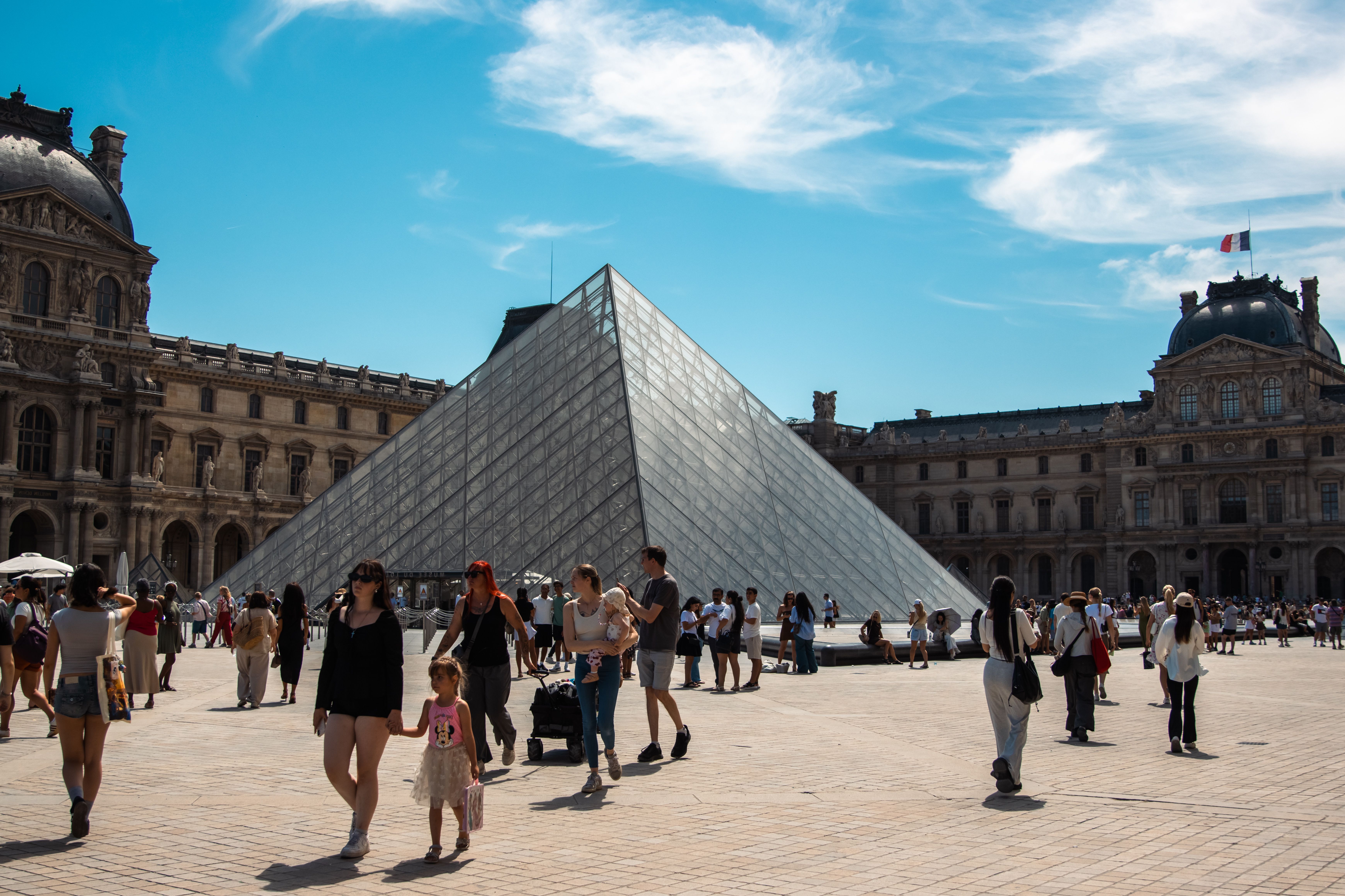 Tourists gather at the Louvre Pyramid in Paris under a clear blue sky.