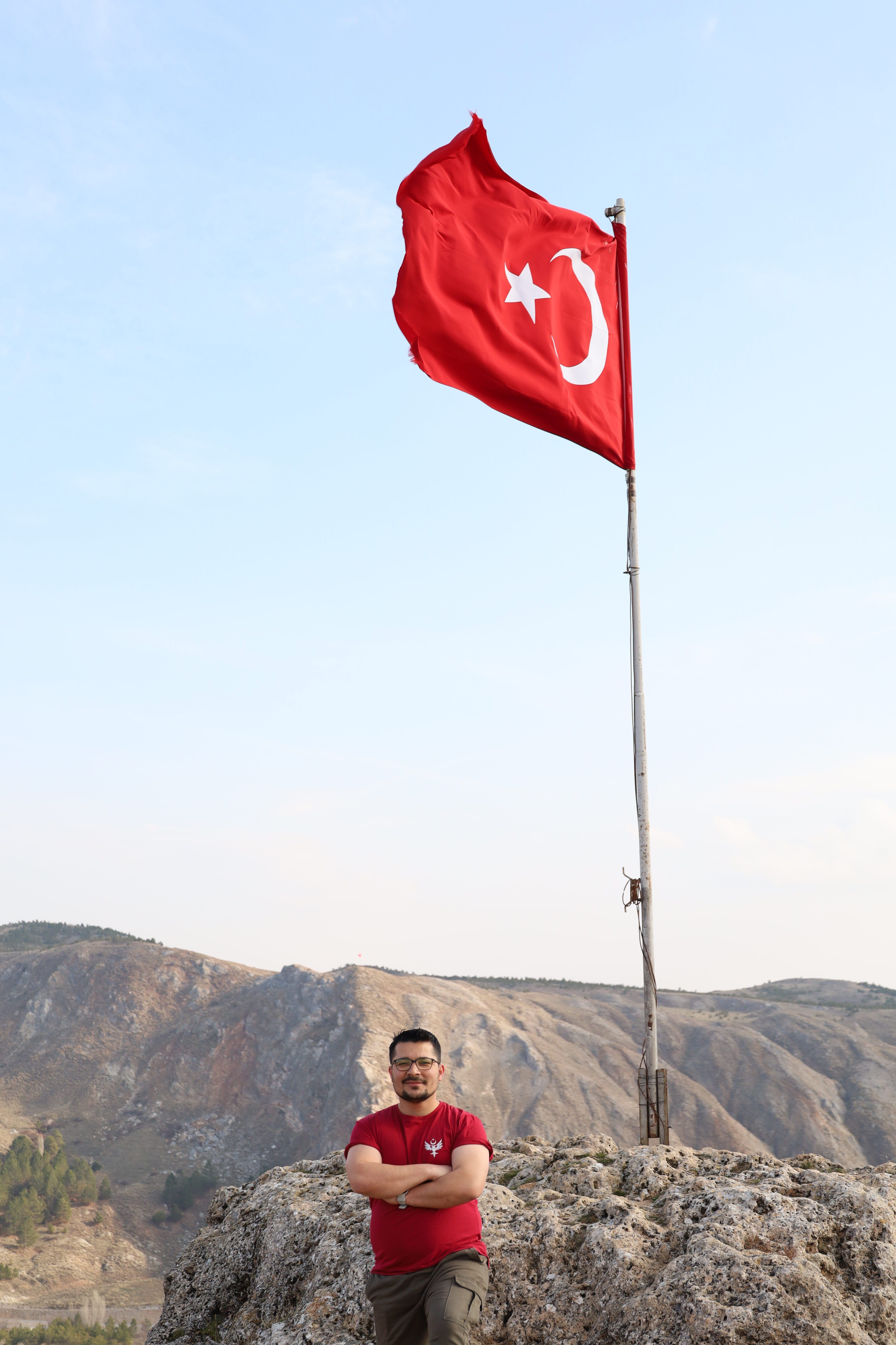 A man poses next to a waving Turkish flag on a rocky hill with a mountainous backdrop.