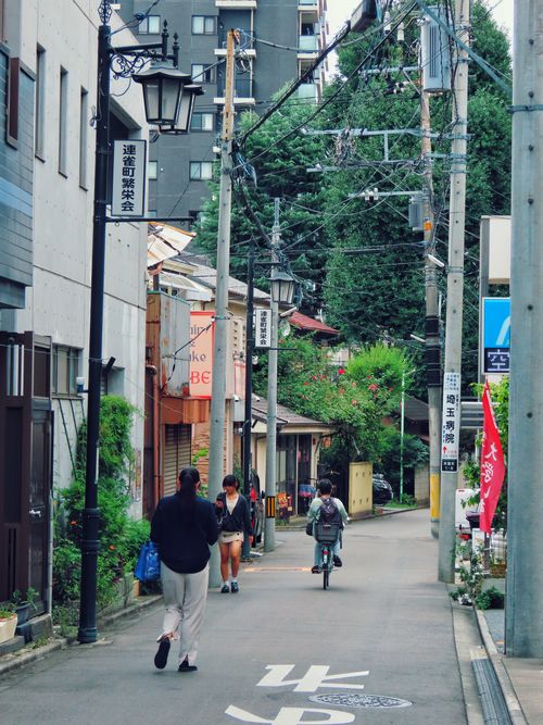 Free A bustling narrow Japanese street with people walking and a cyclist, surrounded by urban buildings and greenery. Stock Photo