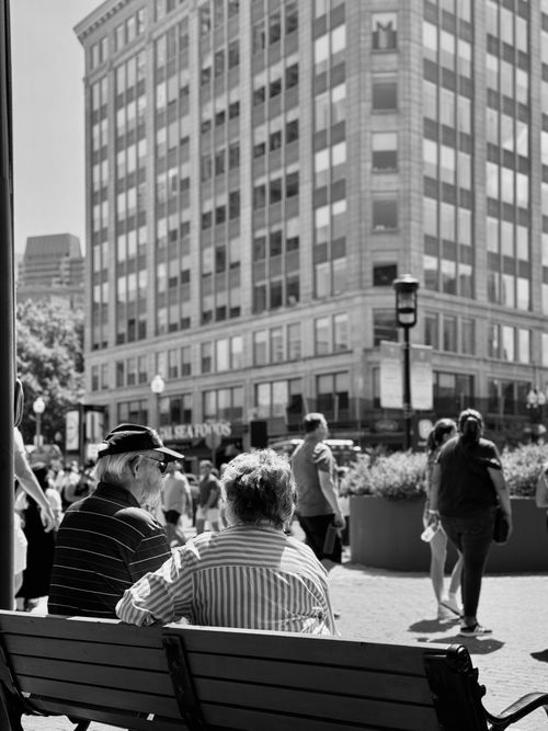 Free An elderly couple enjoys a sunny day sitting on a bench in downtown Boston as pedestrians pass by. Stock Photo