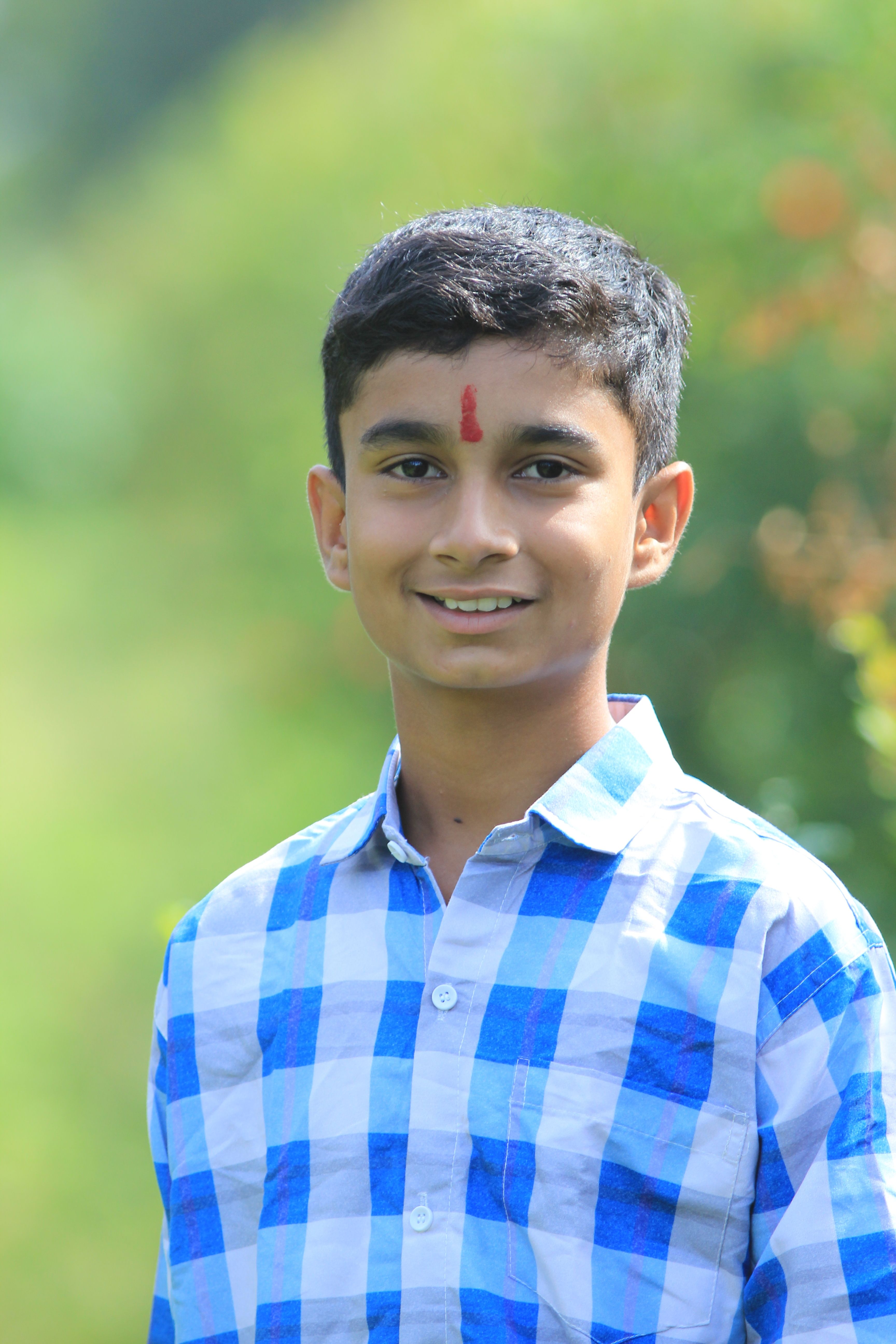 Portrait of a smiling teenager outdoors wearing a blue plaid shirt, marked with red tilak.