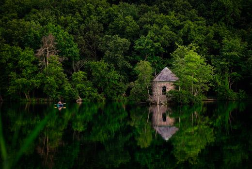 A stone cabin by a tranquil lake surrounded by lush greenery and reflections.