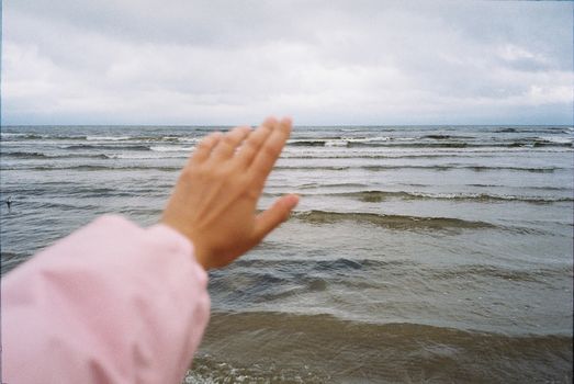 A person's hand waving toward the calm sea under a cloudy sky.