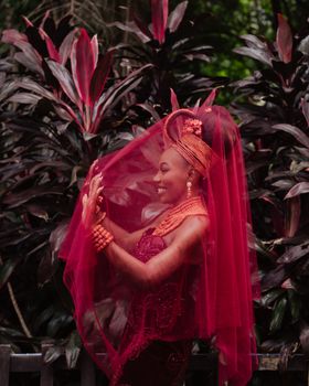 Vibrant Nigerian bride in traditional attire surrounded by lush foliage.