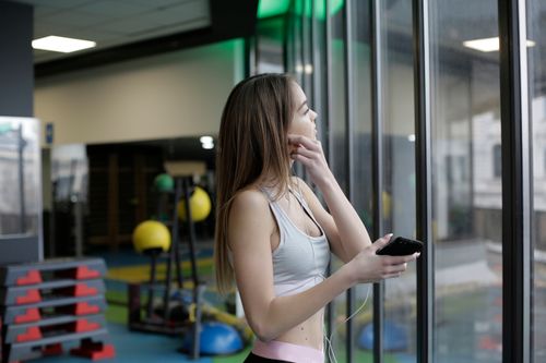 Free Woman in gym wear looking out window, holding a smartphone and earphones. Stock Photo
