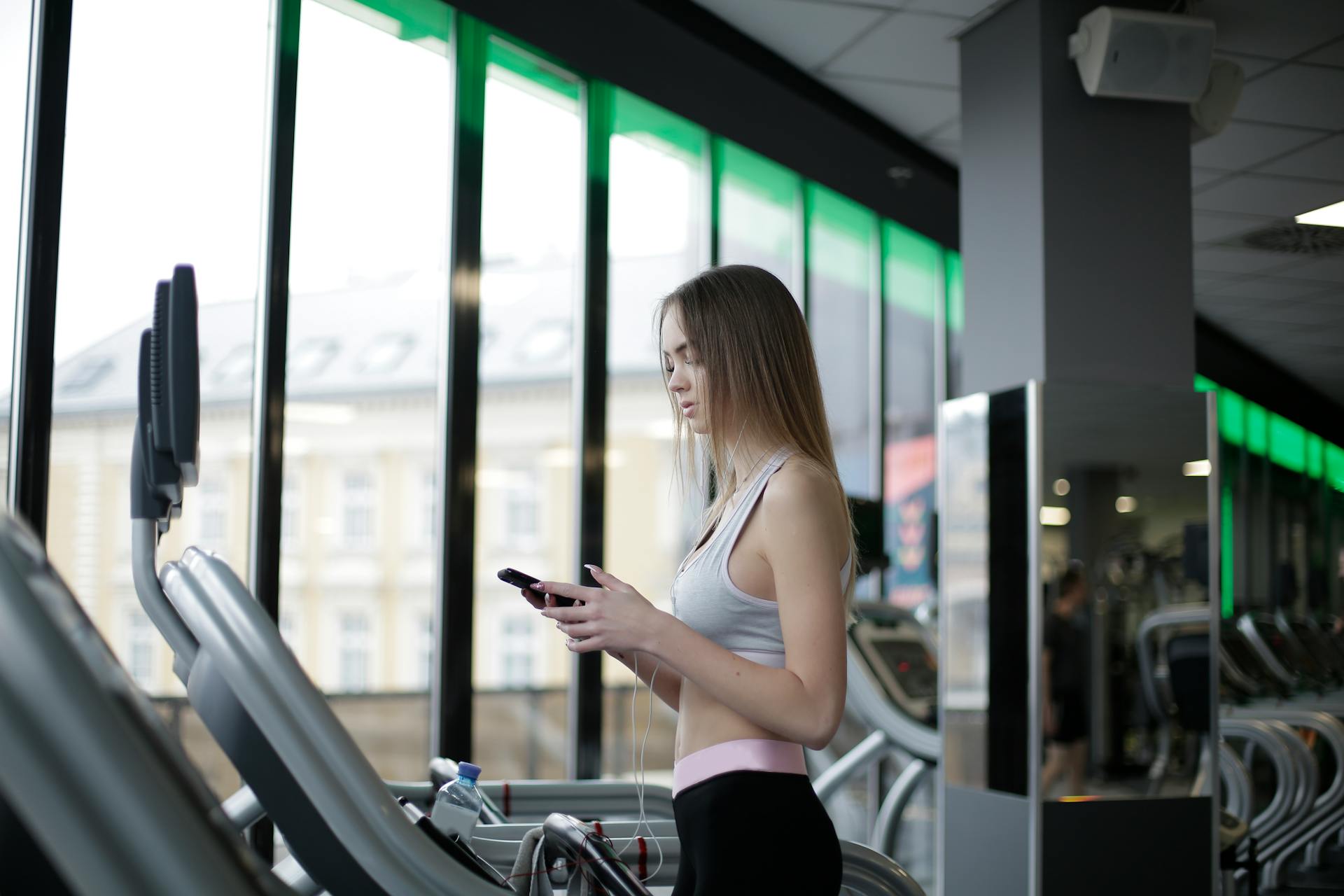 Side view of slim young female athlete in sportswear standing with smartphone and training on treadmill in spacious modern fitness center