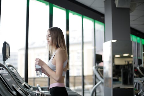 Free Side view of slim young female athlete in activewear standing on treadmill with bottle of fresh water while resting during training in modern light gym Stock Photo