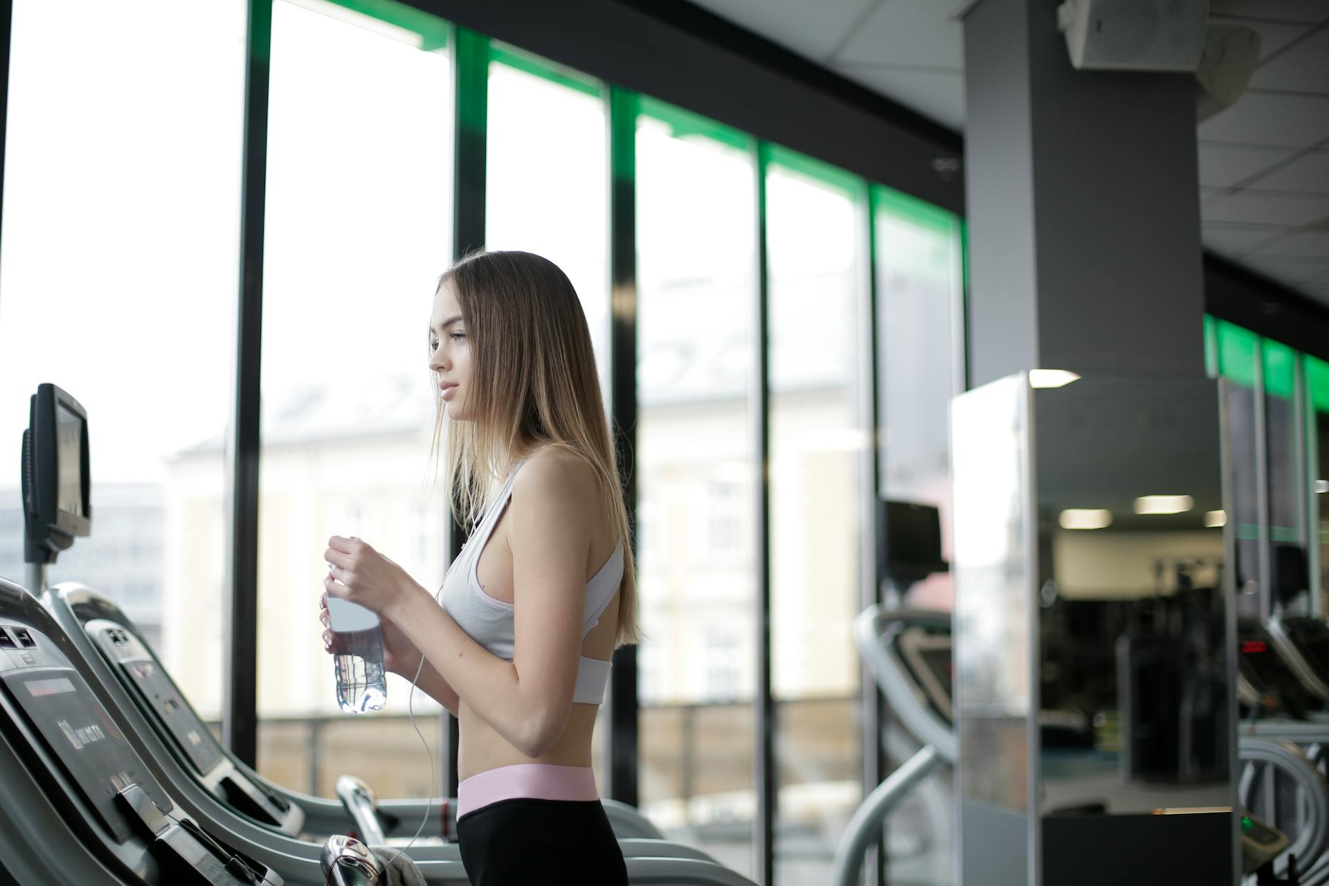 Side view of slim young female athlete in activewear standing on treadmill with bottle of fresh water while resting during training in modern light gym