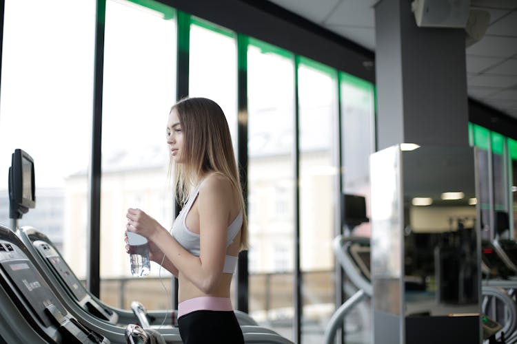 Sporty Young Female Athlete Training On Treadmill With Bottle Of Water In Gym