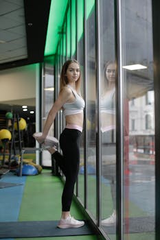 Young woman stretching in a modern gym, reflected in a large window, promoting health and fitness.