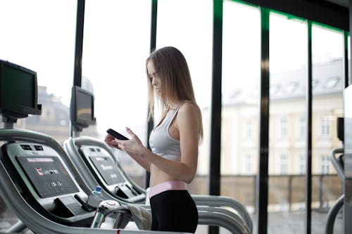 Side view of slim young female athlete in activewear using smartphone while training on treadmill in spacious modern fitness club
