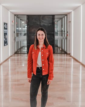 Casual indoor portrait of a stylish woman standing in a bright hallway wearing a red jacket.