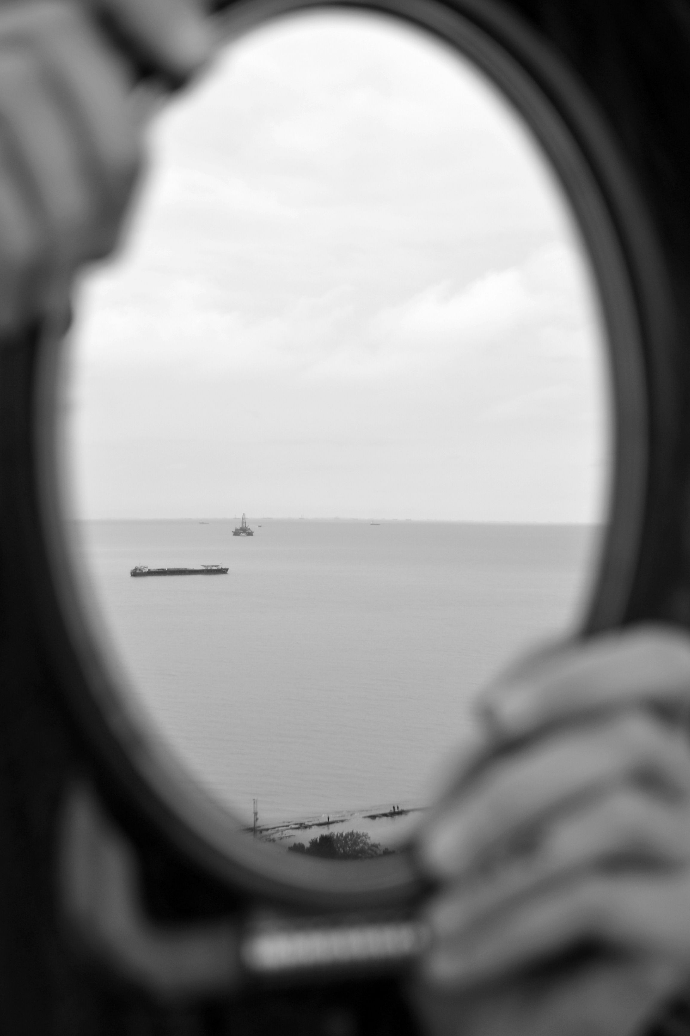 A serene black and white view of the ocean through a round porthole with hands holding it.