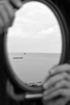A serene black and white view of the ocean through a round porthole with hands holding it.