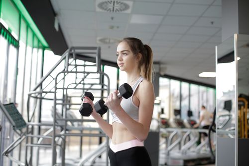 Free Strong young female athlete in activewear doing bicep curls with dumbbells while training in light spacious modern gym and looking away thoughtfully Stock Photo