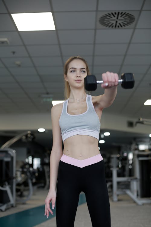 From below view of young slim female athlete in activewear doing dumbbell front raises while training alone in modern spacious fitness center