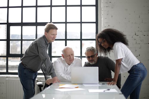 Concentrated multiethnic group of colleagues sitting and standing around laptop on table and discussing project together in light creative workspace