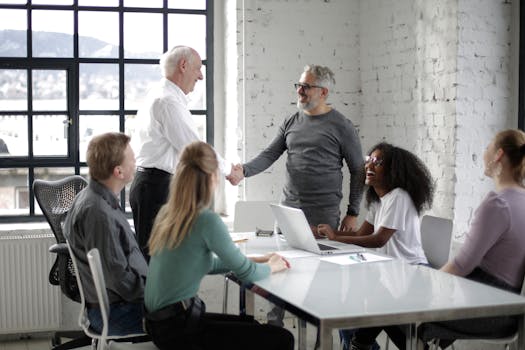 Cheerful male colleagues shaking hands while discussing business ideas with group of multiethnic coworkers gathering around table with gadgets and documents in modern light workspace