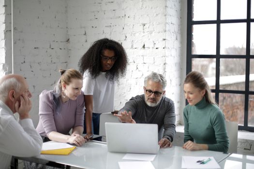 Group of serious multiracial colleagues sitting together at table near laptop and discussing details of project while working in spacious modern workspace