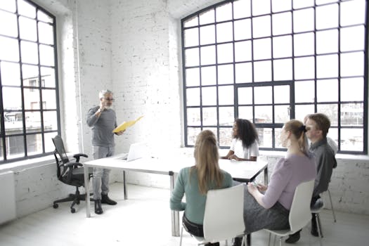 Side view of focused multiethnic colleagues listening middle aged speaker while gathering at table in spacious workspace against wide windows