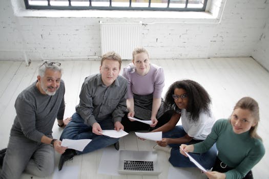 A diverse team of adults collaborating in a modern office, working on documents around a laptop.