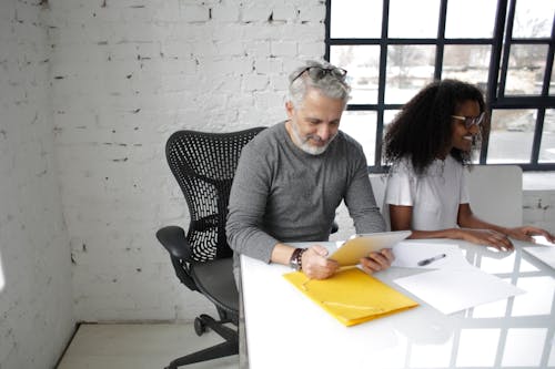 Free Smiling mature male executive surfing tablet while sitting near African American female colleague in eyeglasses working together in creative office Stock Photo
