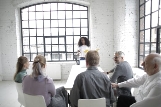 Diverse group discussing ideas in a bright conference room with large windows and a white brick interior.